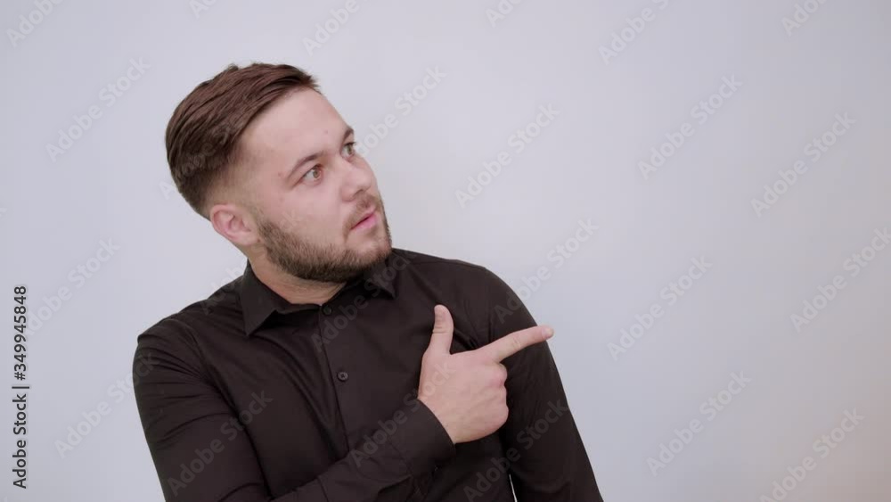 Young Bearded Dark Haired Man In Black Stylish Shirt On White Background, Confused Male Shows Direction Forefinger. The Concept Of Guilt, Slander. Human Relations Young Bearded Dark Haired Man In Black Stylish Shirt On White Background, Confused Male Shows Direction Forefinger. The Concept Of Guilt, Slander. Human Relations