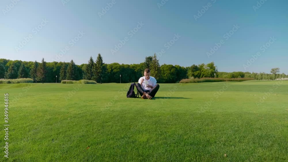 Businessman is eating his vegetarian lunch with eco-friendly packaging. Man sits in park on green grass and eats salad with tomatoes, cucumbers and lettuce in paper package with a wooden fork Businessman is eating his vegetarian lunch with eco-friendly packaging. Man sits in park on green grass and eats salad with tomatoes, cucumbers and lettuce in paper package with a wooden fork