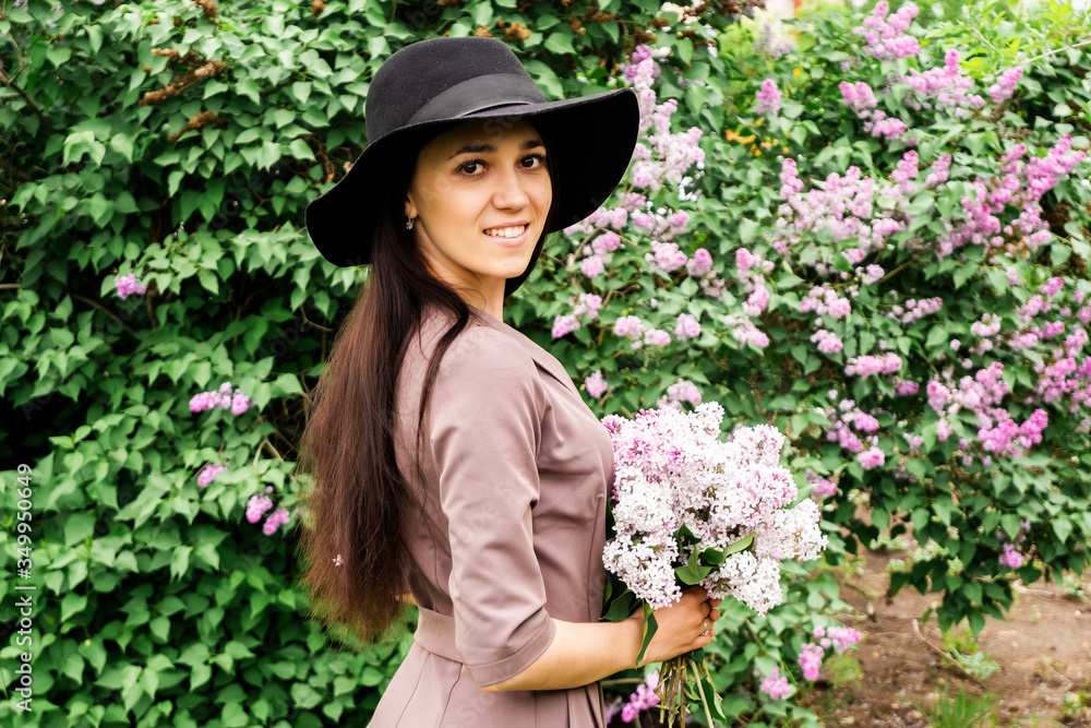 Beautiful young woman with a bouquet of lilacs. Photoshoot of a girl in a lilac. A young brunette in a hat is photographed with a lilac.