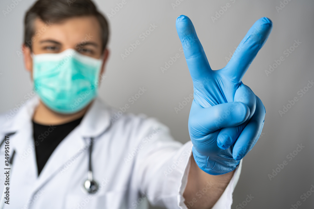 Caucasian man posing as a medical doctor in white overall or scrub with stethoscope around a neck and face mask. Showing peace sign with a hand in blue protective glove. Caucasian man posing as a medical doctor in white overall or scrub with stethoscope around a neck and face mask. Showing peace sign with a hand in blue protective glove.