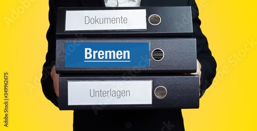 Bremen. Anwalt im Anzug trägt einen Stapel Ordner. 3 Aktenordner. Blaue Beschriftung mit Text. Hintergrund gelb. Recht, Gesetz, Rechtsprechung