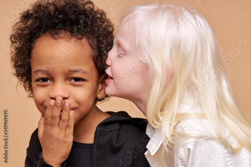 albino girl with pale skin and white hair color tells a secret in the ear of multiracial african boy. cute boy with black skin attentively listen to secret. isolated photo