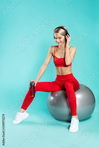 Perfect song for workout. Young and happy sporty blonde woman in headphones sitting on fitness ball and smiling against blue background. Full length