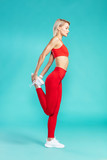 Stretching legs. Side view of young attractive blonde woman in red sportswear warming up before workout while standing against blue background