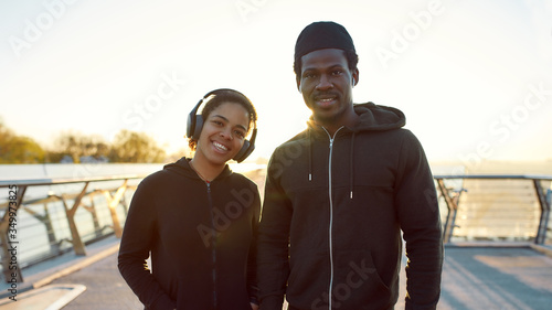 Morning jog. Young happy african couple runners in headphones standing on the bridge outdoors and smiling at camera