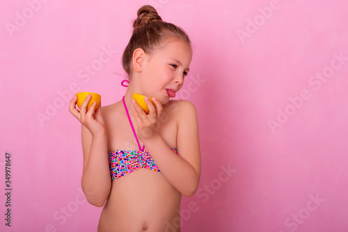 Portrait of pretty child holding slices of lemon. Girl playing with lemon. Little girl eating lemon isolated on pink background, grimacing. Girl shows tongue, has fun.