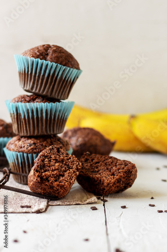 stack of homemade сhocolate muffins with banana in blue paper cups on white, wooden background. Fresh bananas near it.