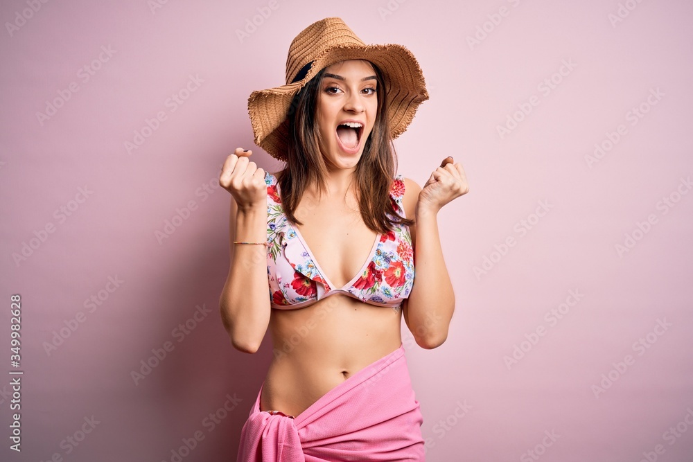 Young beautiful woman on vacation wearing bikini and summer hat over pink background celebrating surprised and amazed for success with arms raised and open eyes. Winner concept. Young beautiful woman on vacation wearing bikini and summer hat over pink background celebrating surprised and amazed for success with arms raised and open eyes. Winner concept.