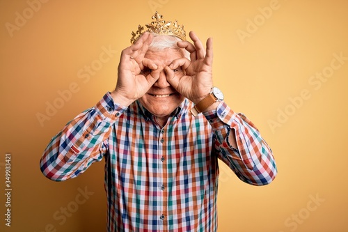 Senior handsome hoary man wearing golden crown of king over isolated yellow background doing ok gesture like binoculars sticking tongue out, eyes looking through fingers. Crazy expression.