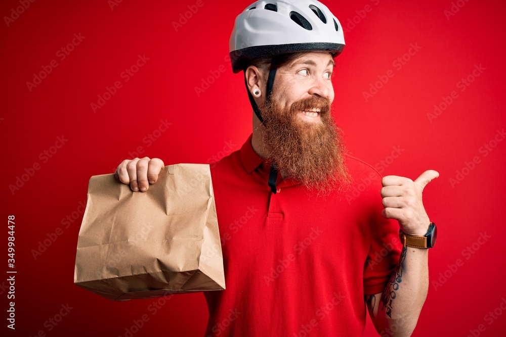 Redhead Irish delivery man with beard wearing bike helmet and holding takeaway paper bag pointing and showing with thumb up to the side with happy face smiling Redhead Irish delivery man with beard wearing bike helmet and holding takeaway paper bag pointing and showing with thumb up to the side with happy face smiling