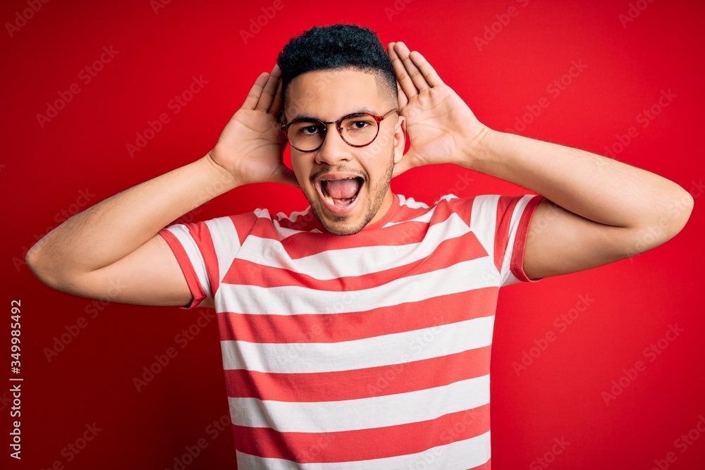 Young handsome man wearing casual striped t-shirt and glasses over isolated red background Smiling cheerful playing peek a boo with hands showing face. Surprised and exited Young handsome man wearing casual striped t-shirt and glasses over isolated red background Smiling cheerful playing peek a boo with hands showing face. Surprised and exited