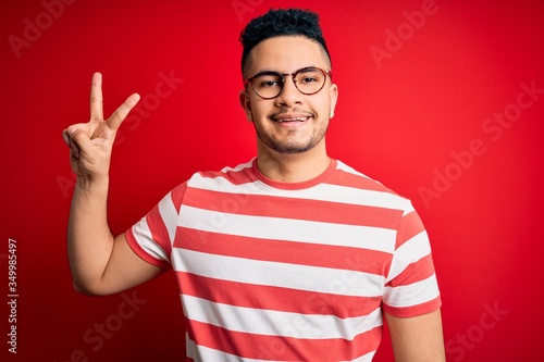 Young handsome man wearing casual striped t-shirt and glasses over isolated red background smiling looking to the camera showing fingers doing victory sign. Number two.