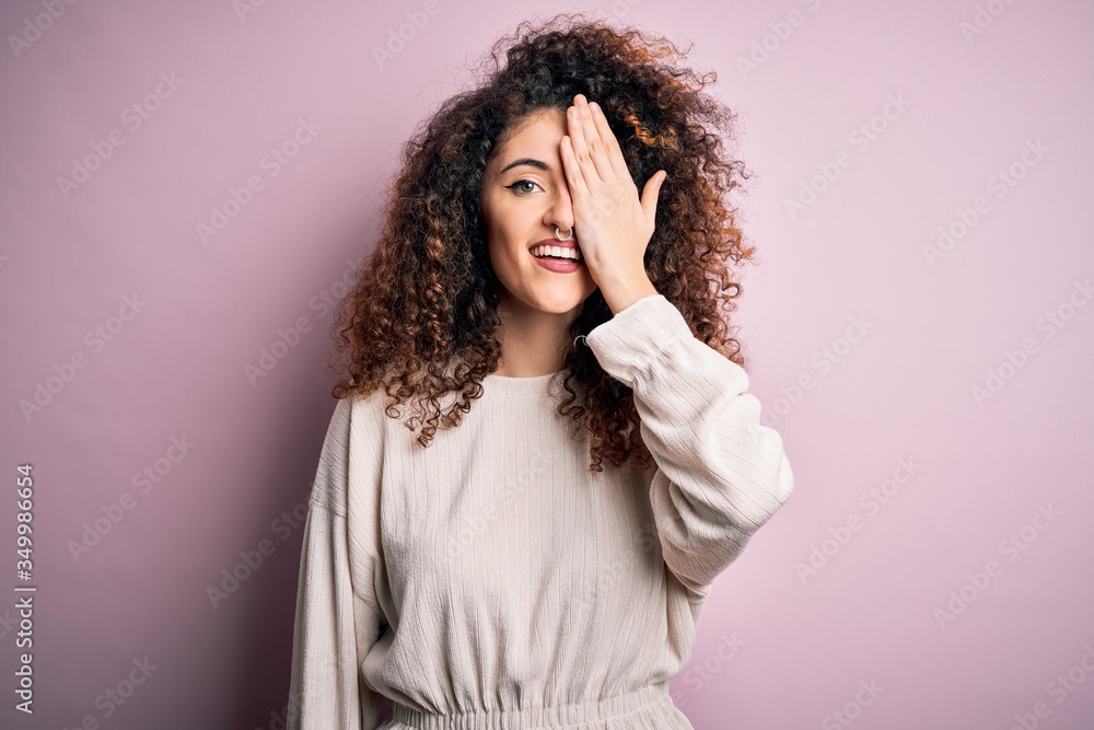 Beautiful woman with curly hair and piercing wearing casual sweater over pink background covering one eye with hand, confident smile on face and surprise emotion. Beautiful woman with curly hair and piercing wearing casual sweater over pink background covering one eye with hand, confident smile on face and surprise emotion.