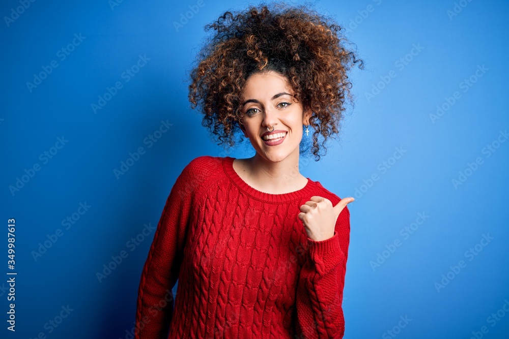 Young beautiful woman with curly hair and piercing wearing casual red sweater smiling with happy face looking and pointing to the side with thumb up. Young beautiful woman with curly hair and piercing wearing casual red sweater smiling with happy face looking and pointing to the side with thumb up.