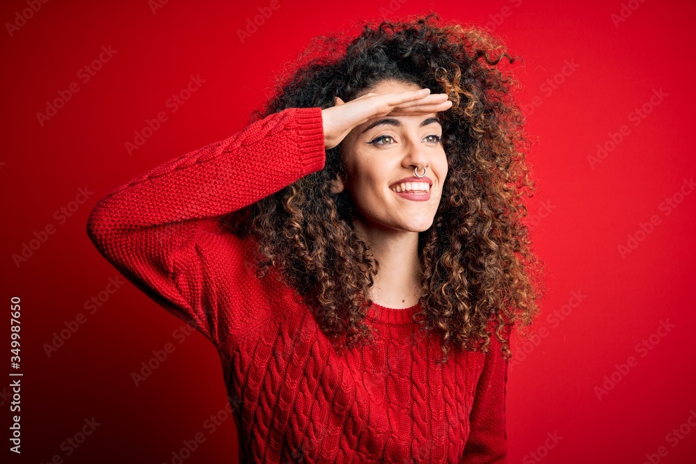 Young beautiful woman with curly hair and piercing wearing casual red sweater very happy and smiling looking far away with hand over head. Searching concept. Young beautiful woman with curly hair and piercing wearing casual red sweater very happy and smiling looking far away with hand over head. Searching concept.