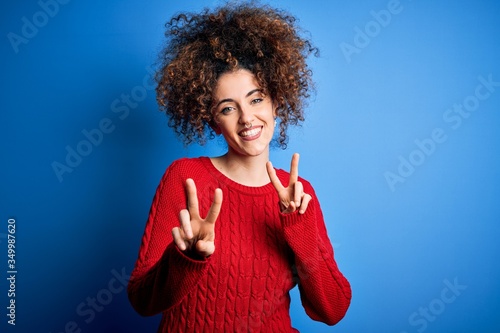 Young beautiful woman with curly hair and piercing wearing casual red sweater smiling looking to the camera showing fingers doing victory sign. Number two.