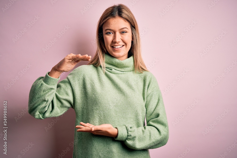 Young beautiful blonde woman wearing winter wool sweater over pink isolated background gesturing with hands showing big and large size sign, measure symbol. Smiling looking at the camera. Measuring Young beautiful blonde woman wearing winter wool sweater over pink isolated background gesturing with hands showing big and large size sign, measure symbol. Smiling looking at the camera. Measuring