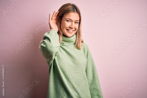Young beautiful blonde woman wearing winter wool sweater over pink isolated background smiling with hand over ear listening an hearing to rumor or gossip. Deafness concept. © Krakenimages.com