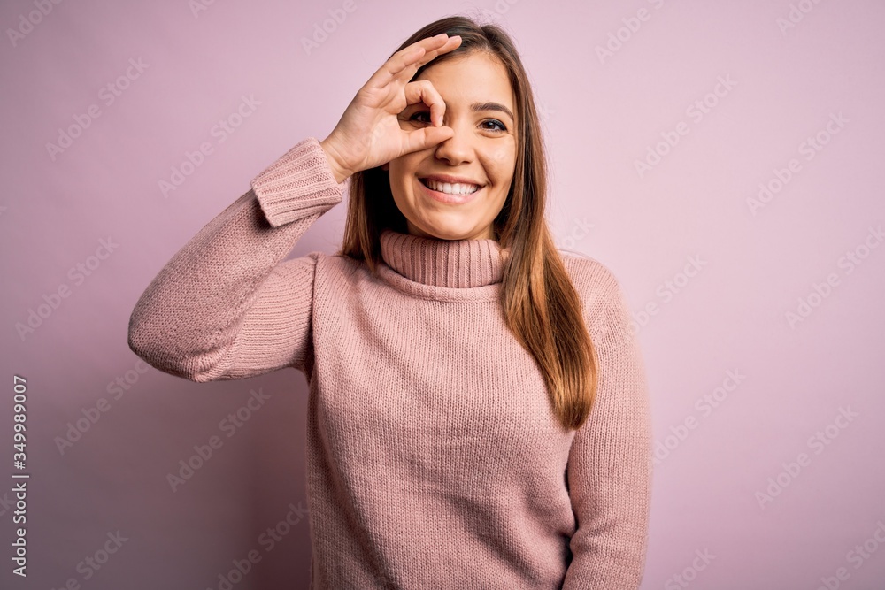 Beautiful young woman wearing turtleneck sweater over pink isolated background doing ok gesture with hand smiling, eye looking through fingers with happy face. Beautiful young woman wearing turtleneck sweater over pink isolated background doing ok gesture with hand smiling, eye looking through fingers with happy face.