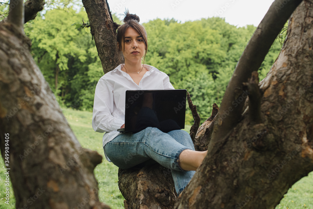 Self-isolation. Quarantine concept. Young girl in a white shirt and blue jeans sitting in nature on an apple tree, working at the computer. Virus. Pandemic. Coronavirus Self-isolation. Quarantine concept. Young girl in a white shirt and blue jeans sitting in nature on an apple tree, working at the computer. Virus. Pandemic. Coronavirus