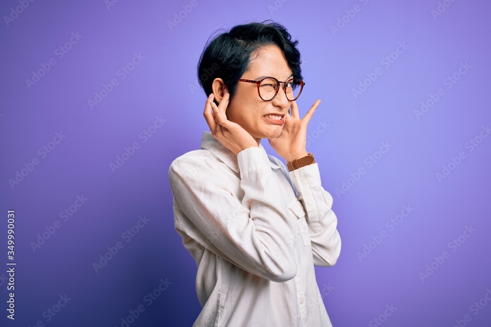 Young beautiful asian girl wearing casual shirt and glasses standing over purple background covering ears with fingers with annoyed expression for the noise of loud music. Deaf concept. Young beautiful asian girl wearing casual shirt and glasses standing over purple background covering ears with fingers with annoyed expression for the noise of loud music. Deaf concept.