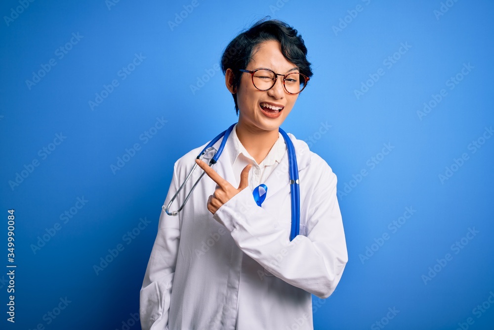 Young beautiful asian doctor girl wearing stethoscope and coat with blue cancer ribbon cheerful with a smile of face pointing with hand and finger up to the side with happy and natural expression