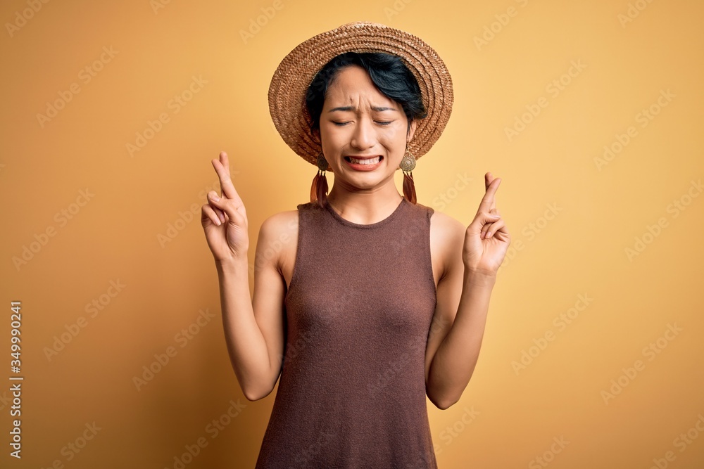 Young beautiful asian girl wearing casual t-shirt and hat over isolated yellow background gesturing finger crossed smiling with hope and eyes closed. Luck and superstitious concept. Young beautiful asian girl wearing casual t-shirt and hat over isolated yellow background gesturing finger crossed smiling with hope and eyes closed. Luck and superstitious concept.