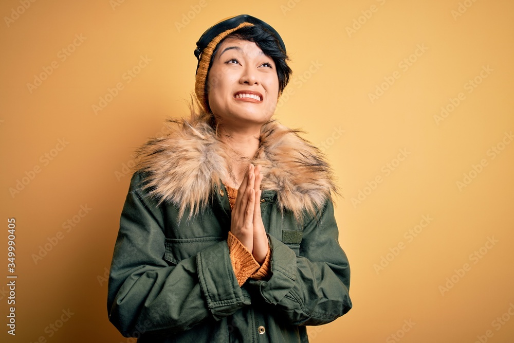 Beautiful asian skier girl wearing snow sportswear using ski goggles over yellow background begging and praying with hands together with hope expression on face very emotional and worried. Begging.