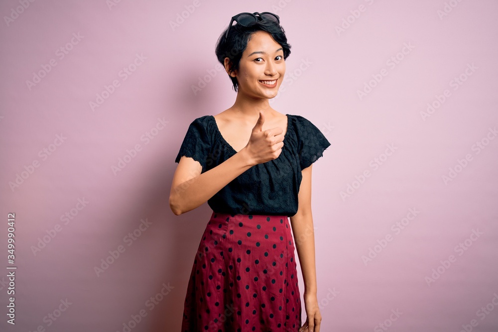 Young beautiful asian girl wearing casual dress standing over isolated pink background doing happy thumbs up gesture with hand. Approving expression looking at the camera showing success. Young beautiful asian girl wearing casual dress standing over isolated pink background doing happy thumbs up gesture with hand. Approving expression looking at the camera showing success.