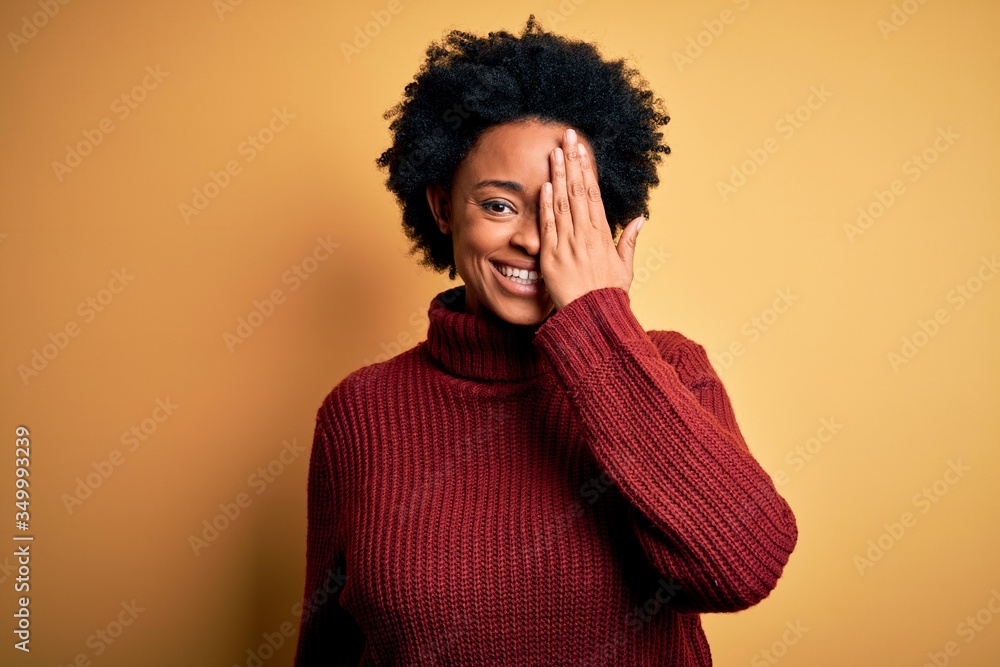 Young beautiful African American afro woman with curly hair wearing casual turtleneck sweater covering one eye with hand, confident smile on face and surprise emotion. Young beautiful African American afro woman with curly hair wearing casual turtleneck sweater covering one eye with hand, confident smile on face and surprise emotion.