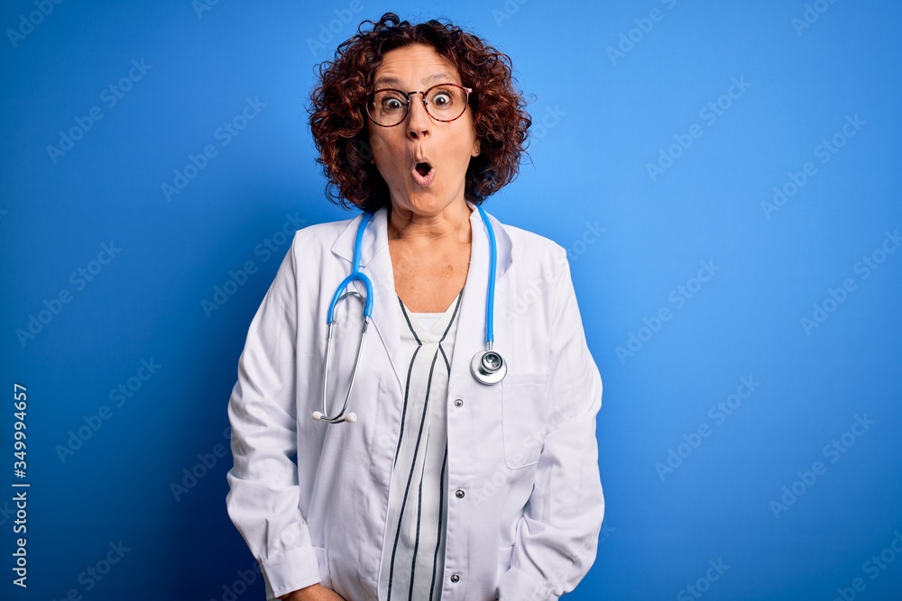 Middle age curly hair doctor woman wearing coat and stethoscope over blue background afraid and shocked with surprise expression, fear and excited face.