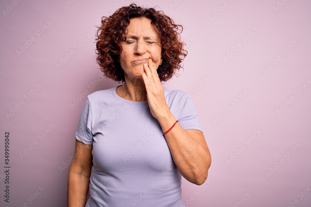 Middle age beautiful curly hair woman wearing casual t-shirt over isolated pink background touching mouth with hand with painful expression because of toothache or dental illness on teeth. Dentist Middle age beautiful curly hair woman wearing casual t-shirt over isolated pink background touching mouth with hand with painful expression because of toothache or dental illness on teeth. Dentist