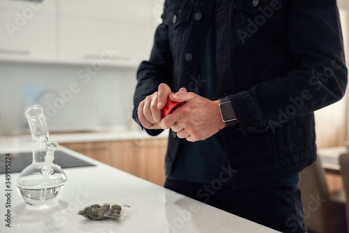 Cropped shot of man using red marijuana grinder, while standing in the kitchen. Glass water pipe or bong, cannabis buds in a plastic bag on the table