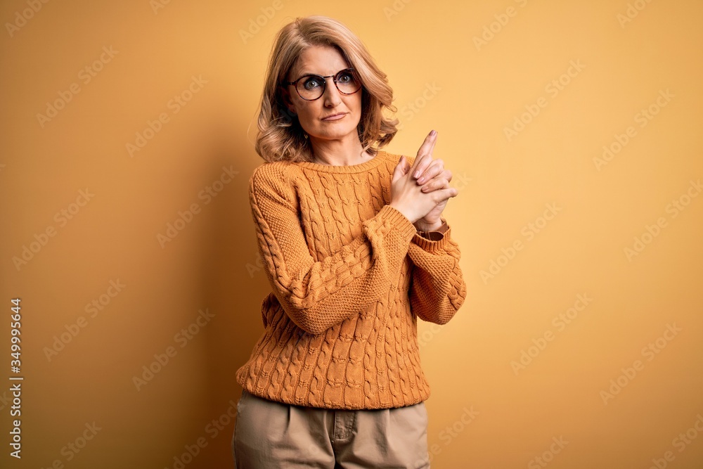 Middle age beautiful blonde woman wearing casual sweater and glasses over yellow background Holding symbolic gun with hand gesture, playing killing shooting weapons, angry face Middle age beautiful blonde woman wearing casual sweater and glasses over yellow background Holding symbolic gun with hand gesture, playing killing shooting weapons, angry face