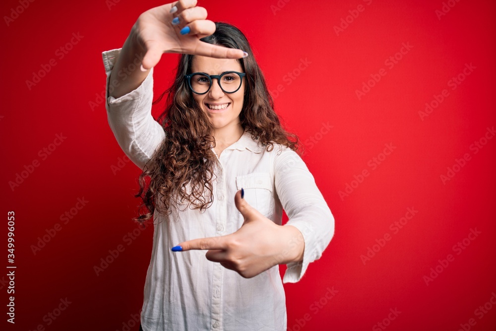 Young beautiful woman with curly hair wearing shirt and glasses over red background smiling making frame with hands and fingers with happy face. Creativity and photography concept. Young beautiful woman with curly hair wearing shirt and glasses over red background smiling making frame with hands and fingers with happy face. Creativity and photography concept.