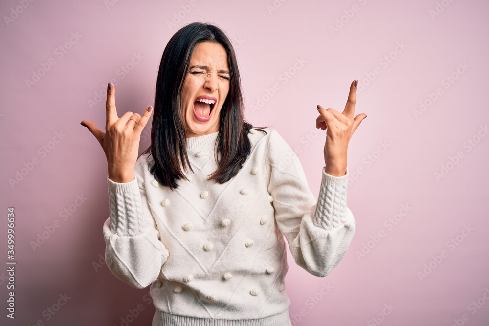 Young brunette woman with blue eyes wearing casual sweater over isolated pink background shouting with crazy expression doing rock symbol with hands up. Music star. Heavy concept. Young brunette woman with blue eyes wearing casual sweater over isolated pink background shouting with crazy expression doing rock symbol with hands up. Music star. Heavy concept.