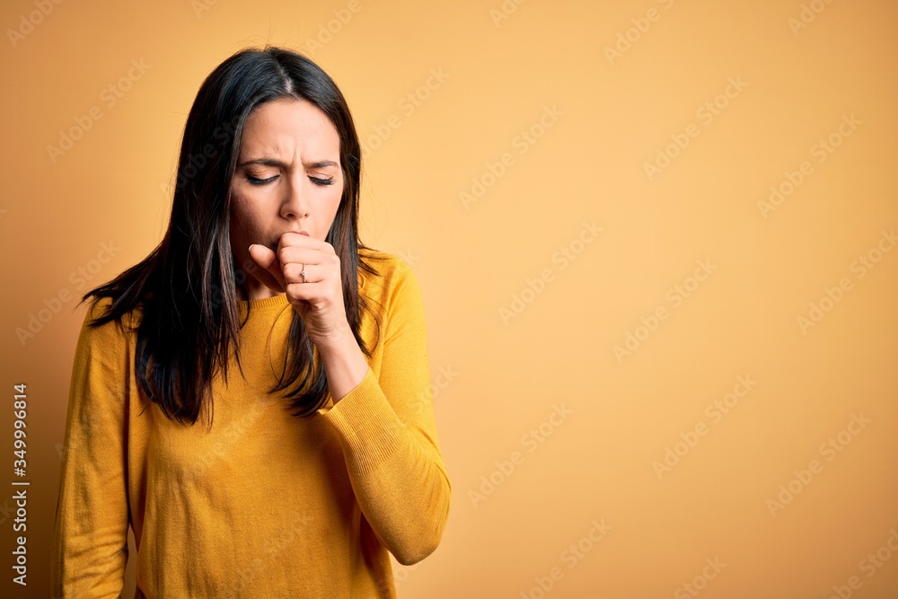 Young brunette woman with blue eyes wearing casual sweater over yellow background feeling unwell and coughing as symptom for cold or bronchitis. Health care concept. Young brunette woman with blue eyes wearing casual sweater over yellow background feeling unwell and coughing as symptom for cold or bronchitis. Health care concept.