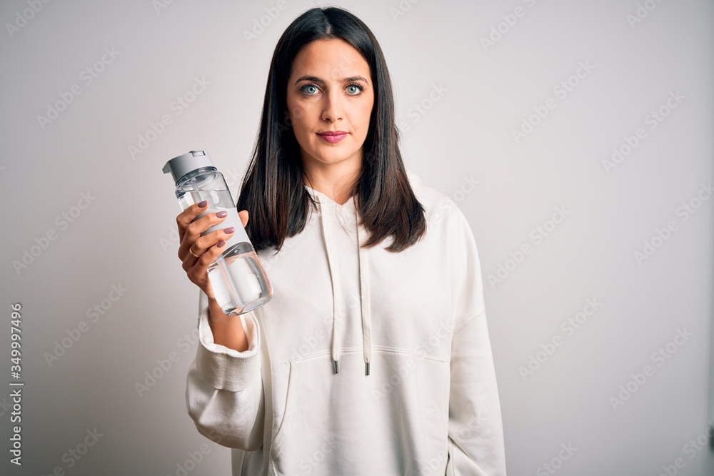 Young brunette sporty woman with blue eyes holding water bottle over white background with a confident expression on smart face thinking serious