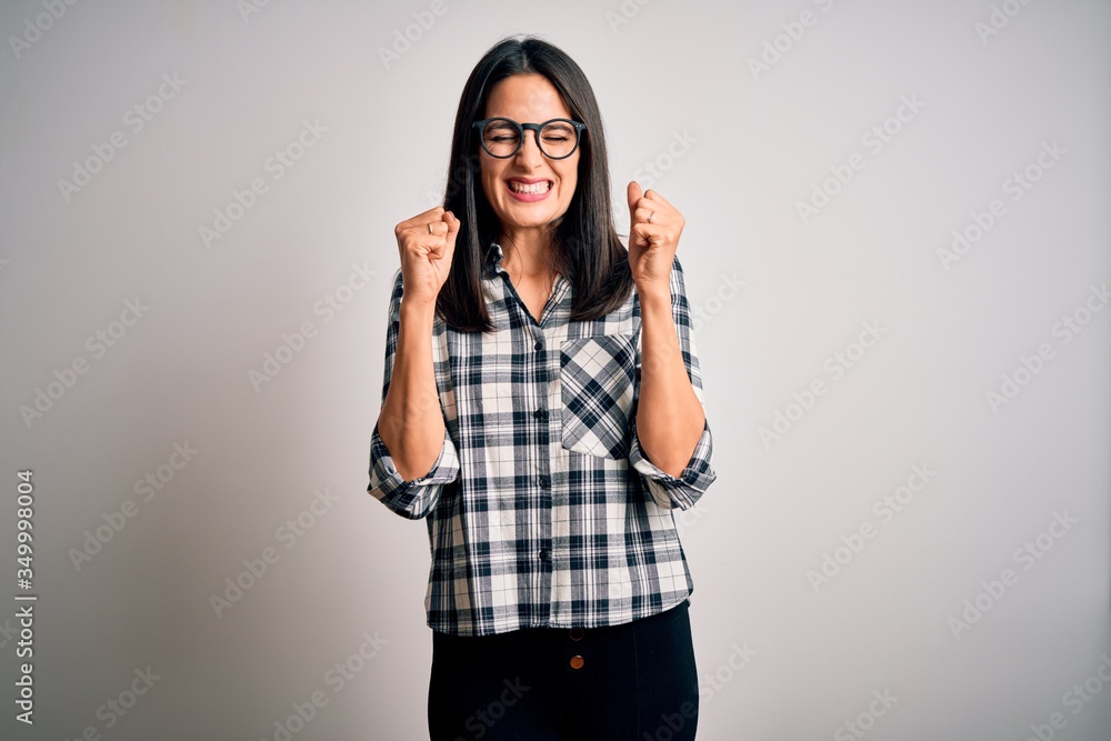 Young brunette woman with blue eyes wearing casual shirt and glasses over white background excited for success with arms raised and eyes closed celebrating victory smiling. Winner concept. Young brunette woman with blue eyes wearing casual shirt and glasses over white background excited for success with arms raised and eyes closed celebrating victory smiling. Winner concept.