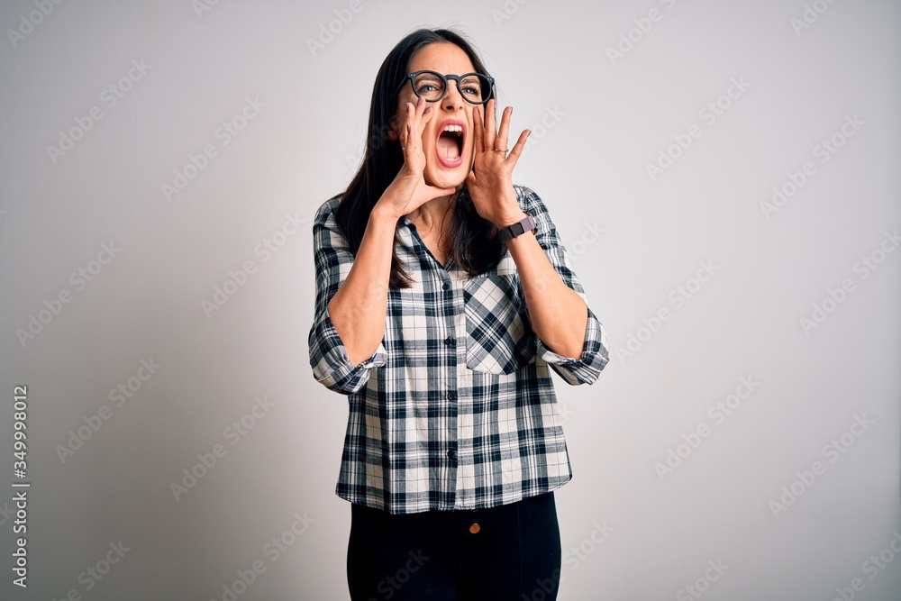 Young brunette woman with blue eyes wearing casual shirt and glasses over white background Shouting angry out loud with hands over mouth Young brunette woman with blue eyes wearing casual shirt and glasses over white background Shouting angry out loud with hands over mouth
