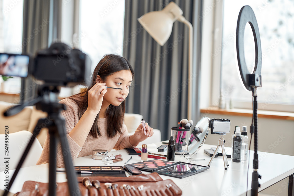 Creative process. Asian female blogger showing her makeup techniques and secrets while recording a tutorial video for her beauty blog using camera at home