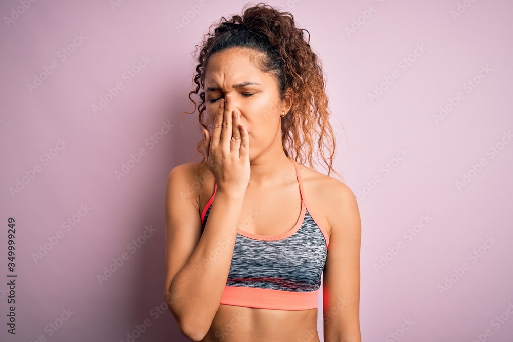 Naklejka premium Beautiful sportswoman with curly hair doing sport wearing sportswear over pink background bored yawning tired covering mouth with hand. Restless and sleepiness.