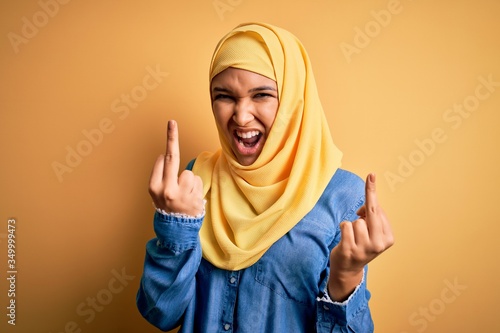 Young beautiful woman with curly hair wearing arab traditional hijab over yellow background Showing middle finger doing fuck you bad expression, provocation and rude attitude. Screaming excited © Krakenimages.com