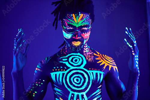 portrait of african man painted in fluorescent paint on face and muscular torso, studio shot with UV light. ethnic prints
