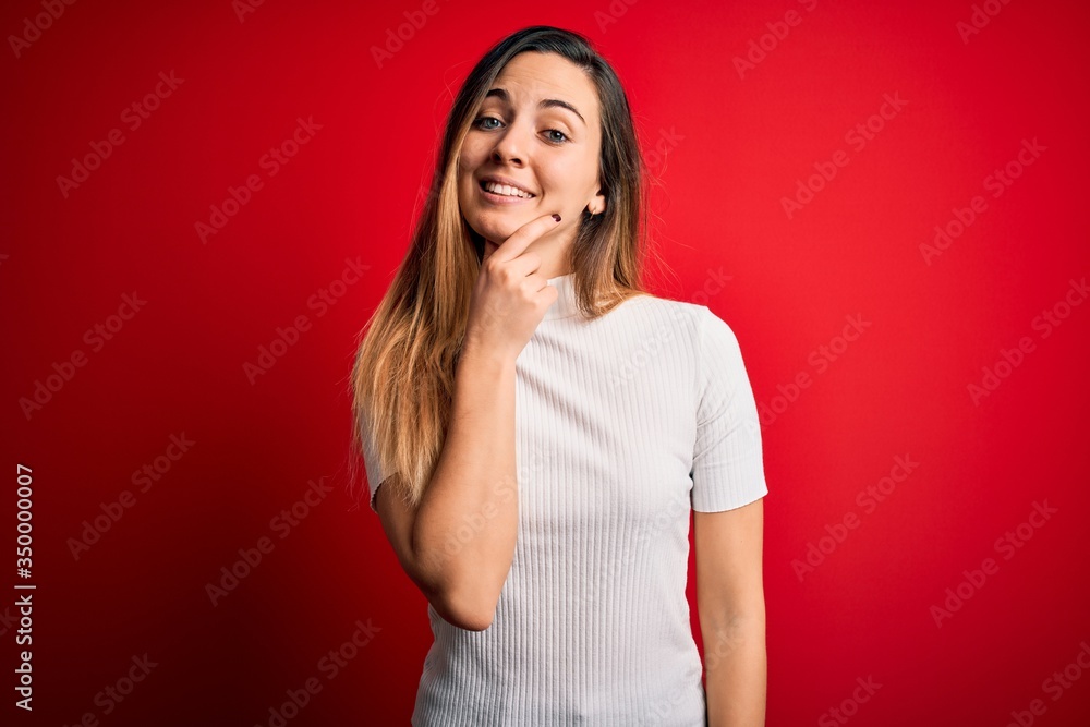 Beautiful blonde woman with blue eyes wearing casual white t-shirt over red background looking confident at the camera smiling with crossed arms and hand raised on chin. Thinking positive. Beautiful blonde woman with blue eyes wearing casual white t-shirt over red background looking confident at the camera smiling with crossed arms and hand raised on chin. Thinking positive.