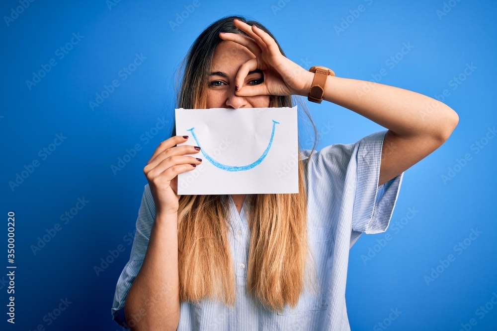 Young beautiful brunette woman holding card with smile illustration on mouth with happy face smiling doing ok sign with hand on eye looking through fingers Young beautiful brunette woman holding card with smile illustration on mouth with happy face smiling doing ok sign with hand on eye looking through fingers