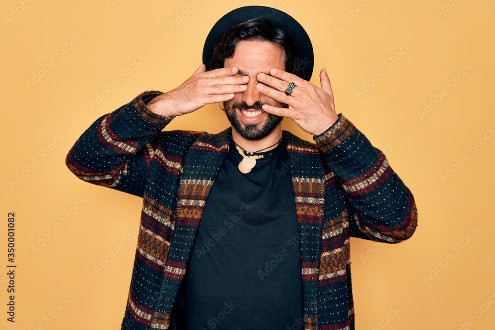 Young handsome hispanic bohemian man wearing hippie style and boho hat covering eyes with hands smiling cheerful and funny. Blind concept. Young handsome hispanic bohemian man wearing hippie style and boho hat covering eyes with hands smiling cheerful and funny. Blind concept.