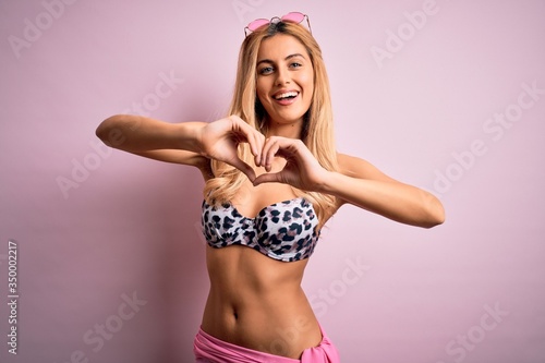 Young beautiful blonde woman on vacation wearing bikini over isolated pink background smiling in love doing heart symbol shape with hands. Romantic concept.