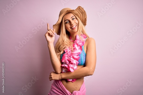 Young beautiful blonde woman on vacation wearing bikini and hat with hawaiian lei flowers with a big smile on face, pointing with hand and finger to the side looking at the camera. © Krakenimages.com