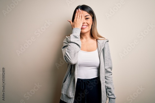 Young beautiful brunette sportswoman wearing sportswoman training over white background covering one eye with hand, confident smile on face and surprise emotion.